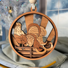 Load image into Gallery viewer, Three-layered Christmas ornament, approximately 4" in diameter, made of multiple hardwood veneers, depicting a fancy rooster sitting on a barnyard fence with sun rays in the background. Made in Virginia by @BelCreativeArts.