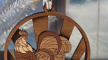Load and play video in Gallery viewer, Video of three-layered Christmas ornament, approximately 4&quot; in diameter, made of multiple hardwood veneers, depicting a fancy rooster sitting on a barnyard fence with sun rays in the background.  Made in Virginia by @BelCreativeArts.
